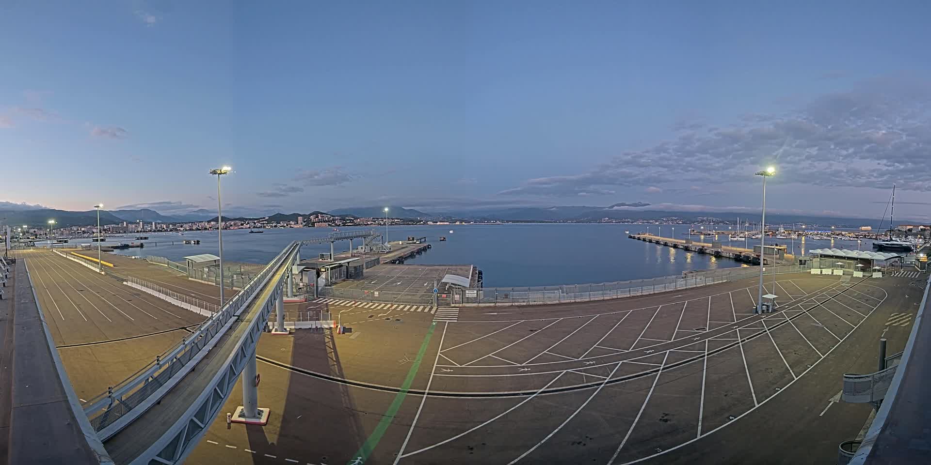 An empty ferry terminal at dusk overlooks a calm body of water with mountains in the distance under a partly cloudy sky.