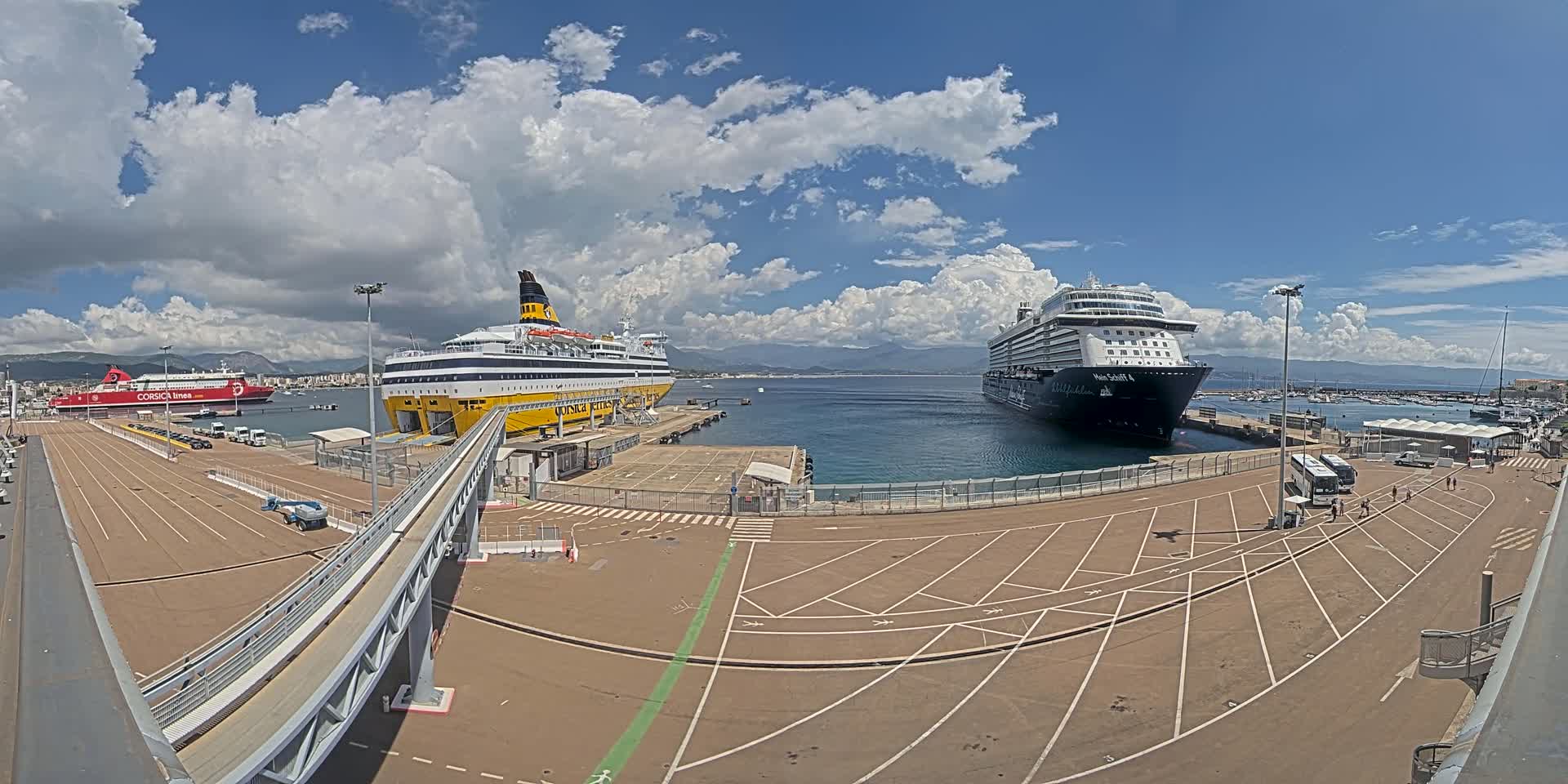 A sunny day at a port shows two large passenger ships docked, one yellow and black, the other dark blue and black, with several vehicles and buses in the parking lot.
