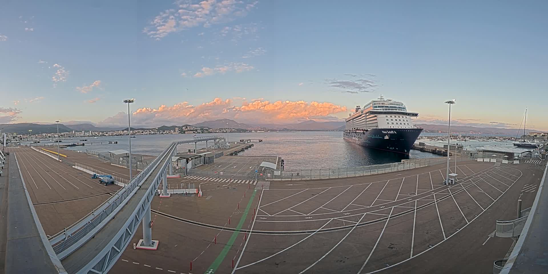 A large cruise ship is docked at a port, with a city and mountains visible in the distance under a partly cloudy, dusky sky.