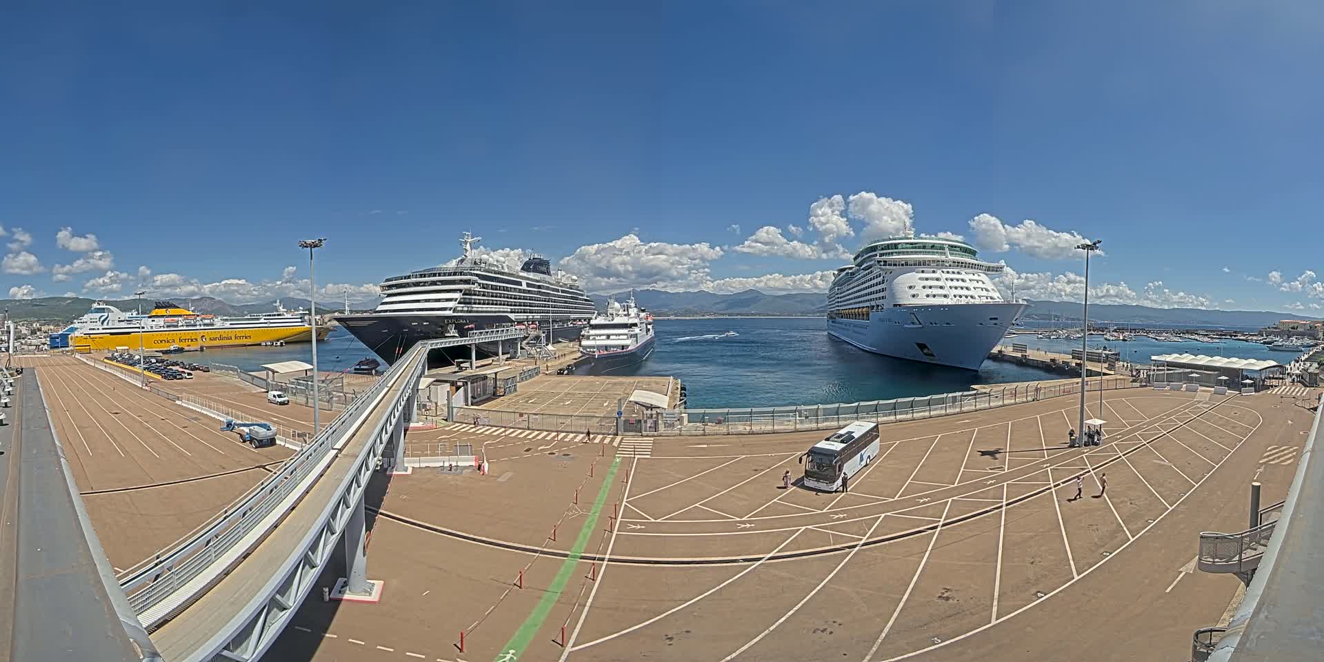 Three large cruise ships and a ferry are docked at a port on a sunny day with partly cloudy skies and mountains in the distance.