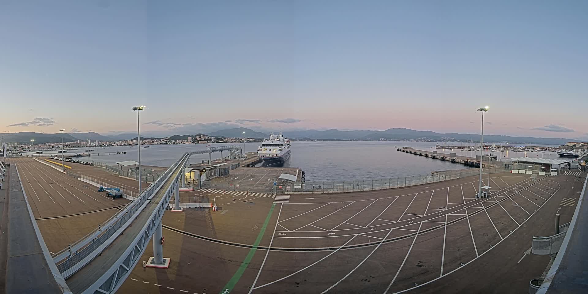 A large passenger ship is docked at a port, with an empty parking lot and a walkway leading to it, under a clear twilight sky.