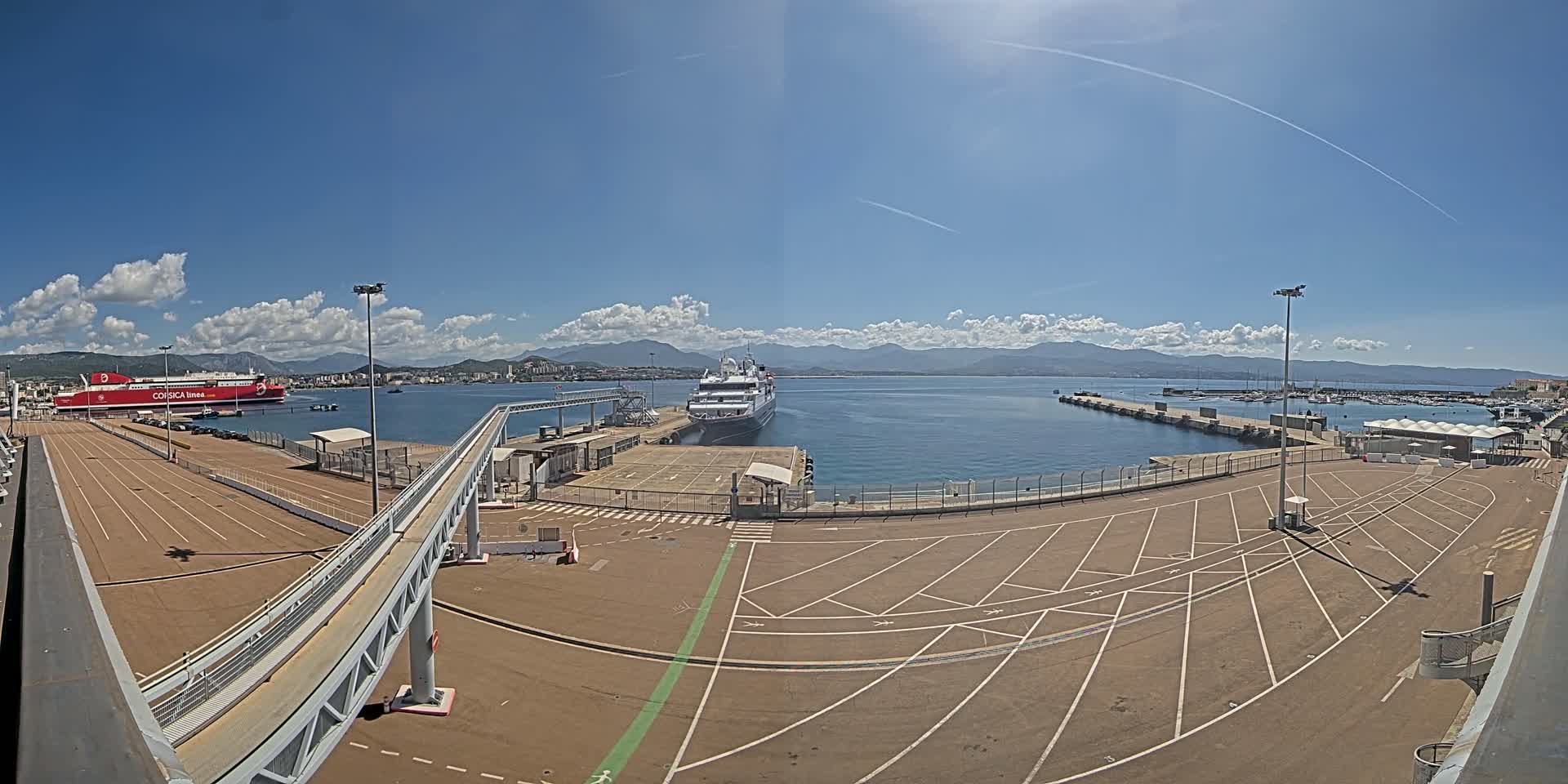 A large empty parking lot at a port, with two docked ships and a distant mountain range under a mostly sunny sky.