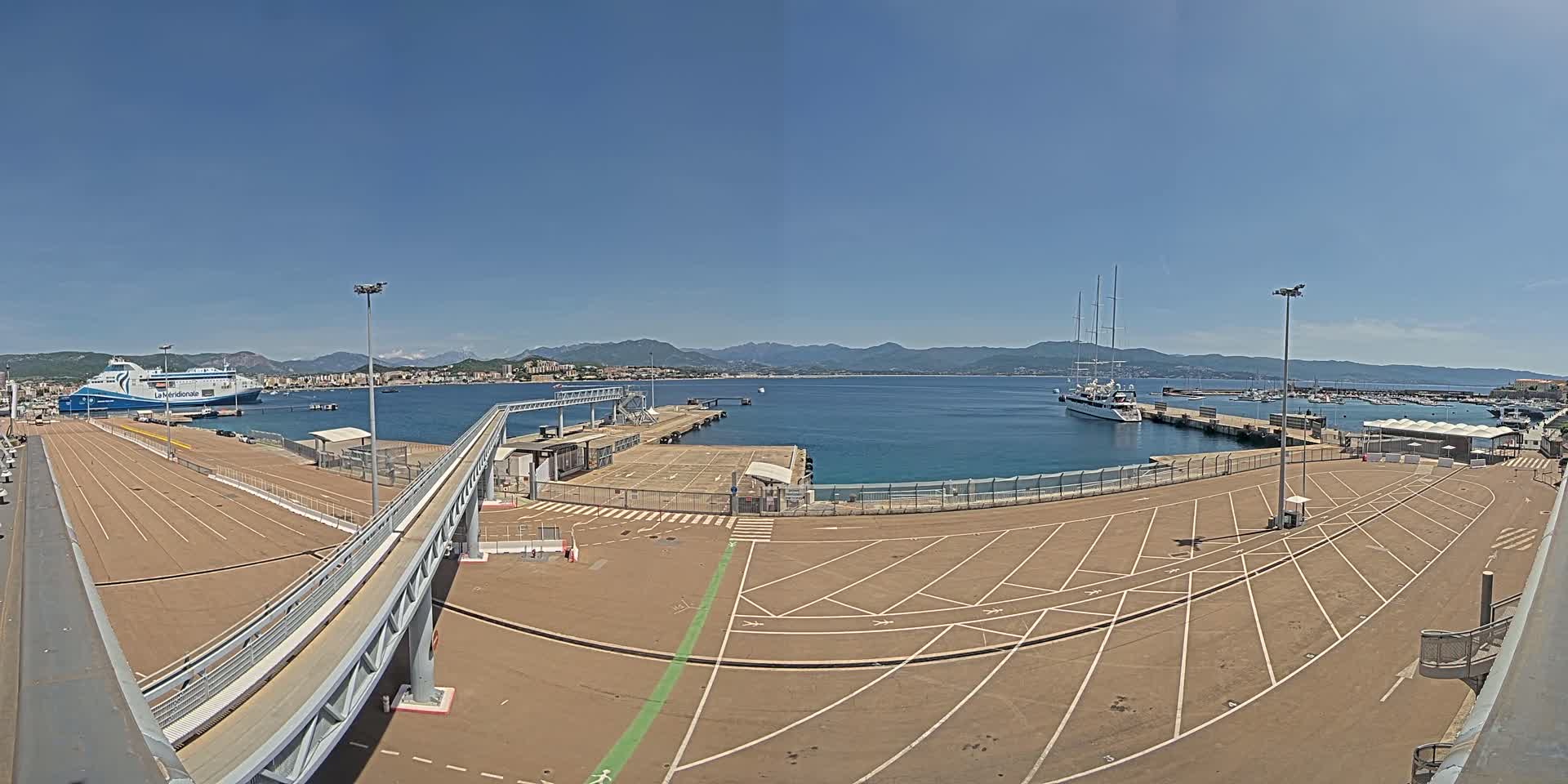 A ferry is docked at a port on a clear, sunny day with mountains in the distance and a large, mostly empty parking lot in the foreground.