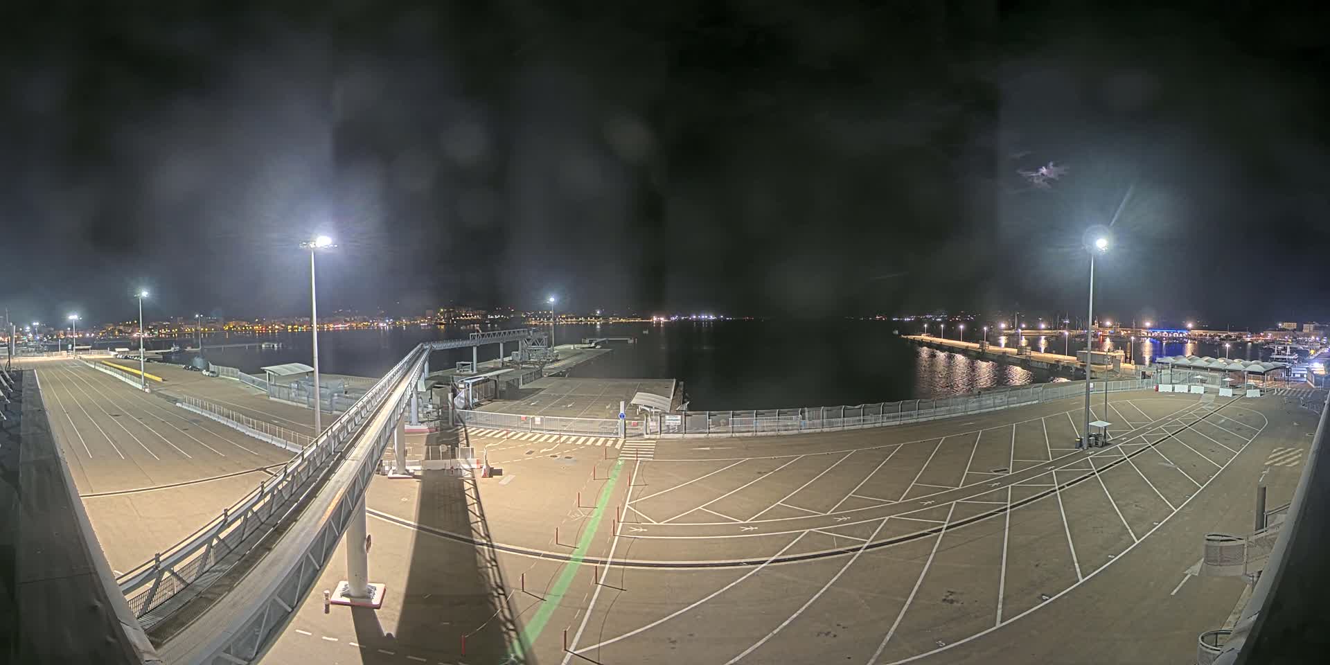 A nighttime view of a mostly empty port area with a long, elevated walkway and a body of water in the background under a dark, cloudy sky.
