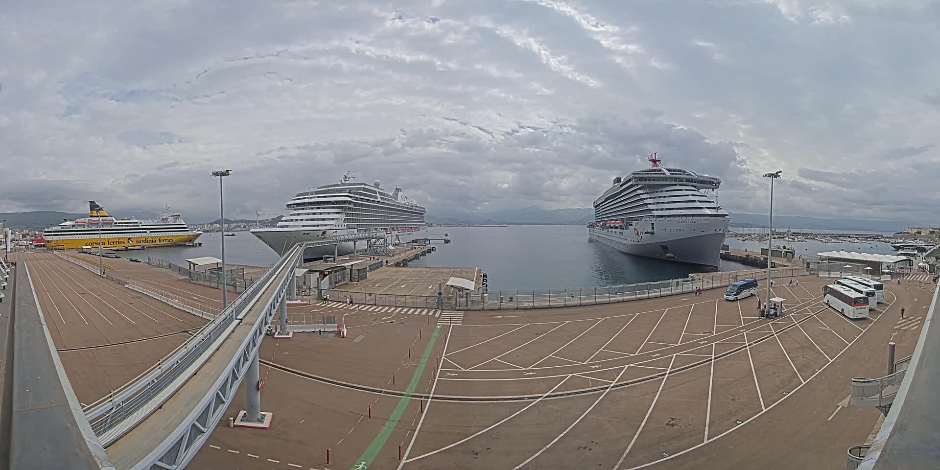A cruise ship and a ferry are docked at a port under a cloudy sky, with several buses and cars parked in the adjacent lot.