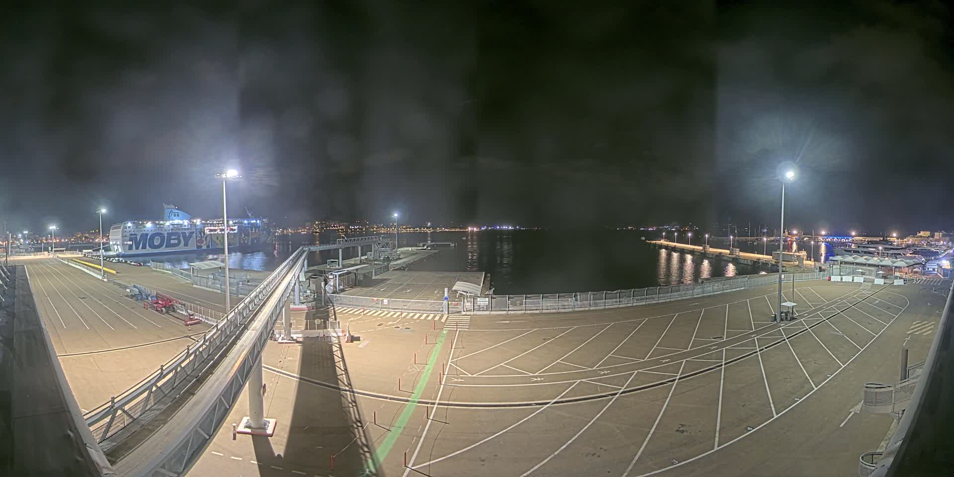 A nighttime view of a ferry docked at a port, with empty parking lots and calm water visible under a cloudy sky.