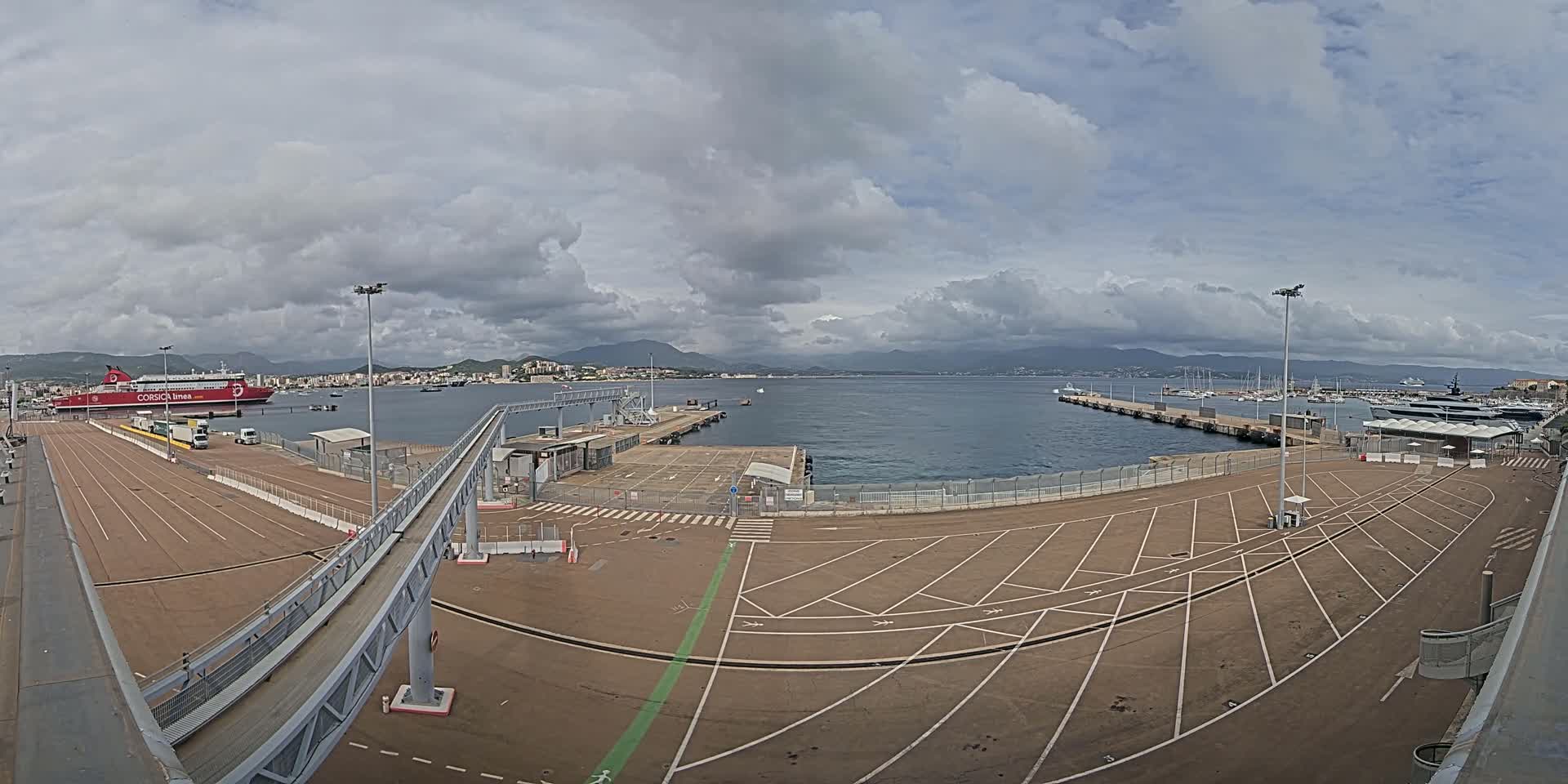 A large empty parking lot at a ferry terminal overlooks a harbor with a docked ferry and numerous boats, under a cloudy sky.