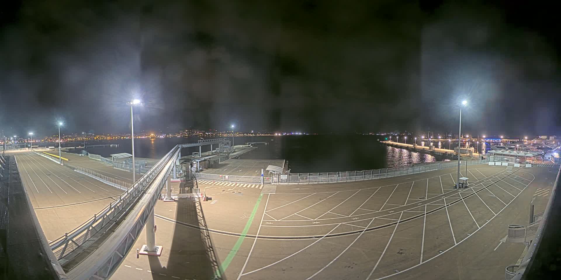 An empty, nighttime ferry terminal with a gangway, parking lot, and a body of water with lights in the distance under a hazy, dark sky.