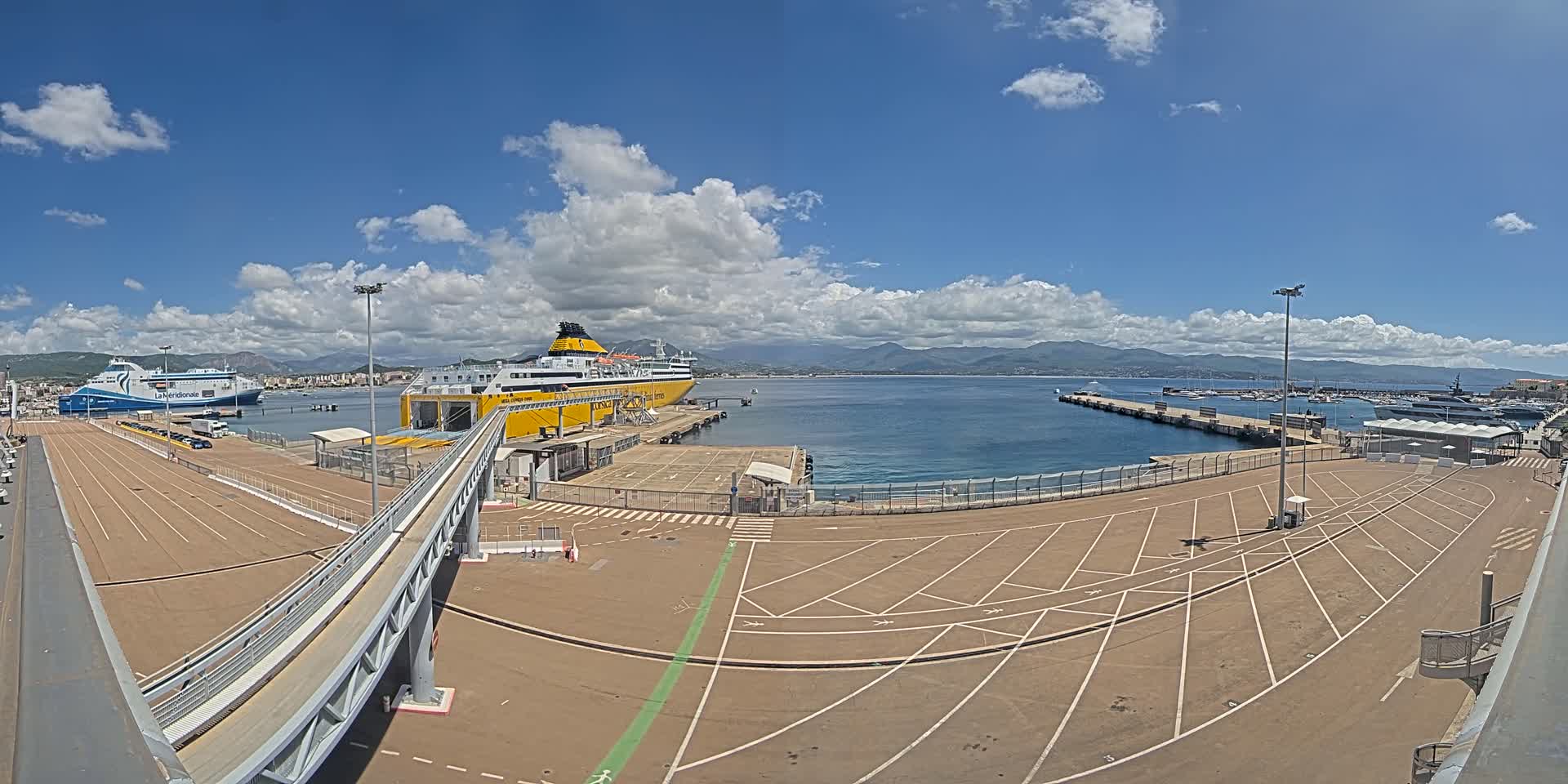 A mostly sunny day at a ferry terminal shows two large ferries docked, one yellow and one blue and white, with a large empty parking lot and a walkway leading to one of the ships.