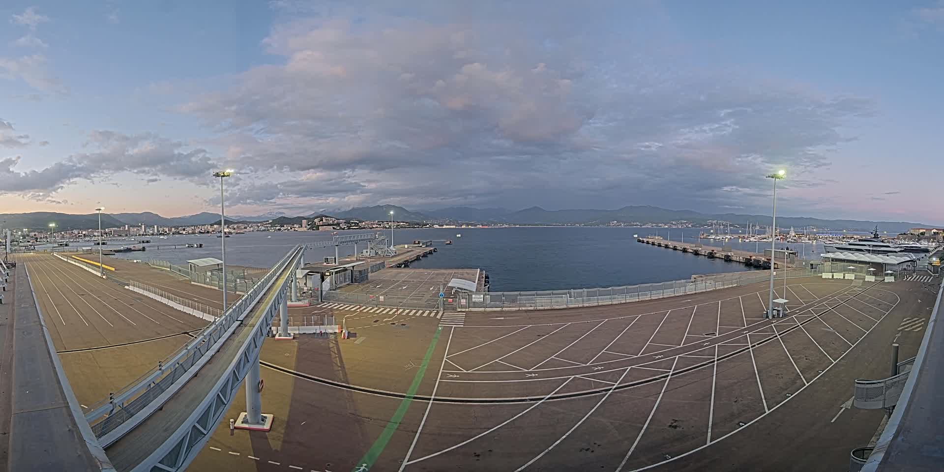 A mostly empty ferry terminal with a walkway leading to a dock overlooks a calm bay and distant city under a cloudy sky.
