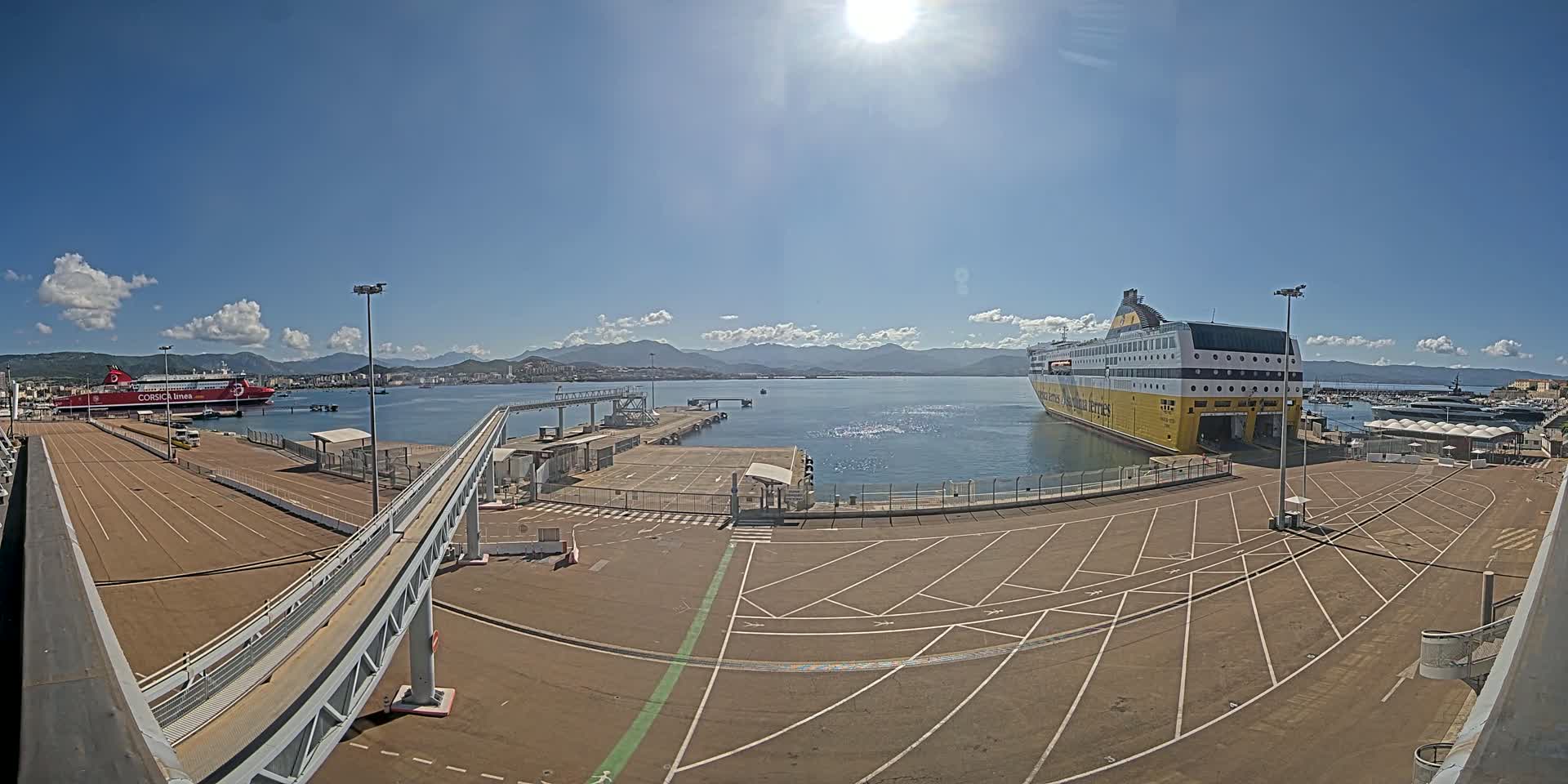 A sunny day at a port shows two ferries docked, one red and one yellow, with a walkway leading to them, and mountains in the background.