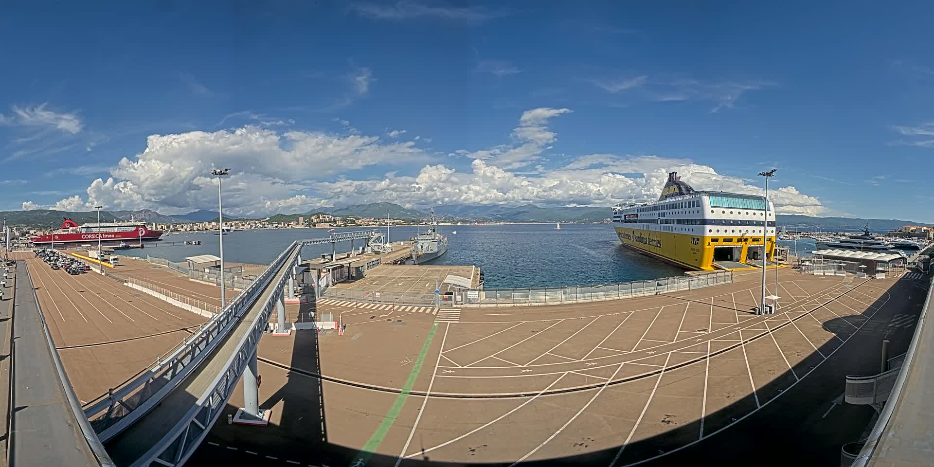 A port with two large ferries docked, a smaller naval vessel, and a mostly empty parking lot under a partly cloudy sky.