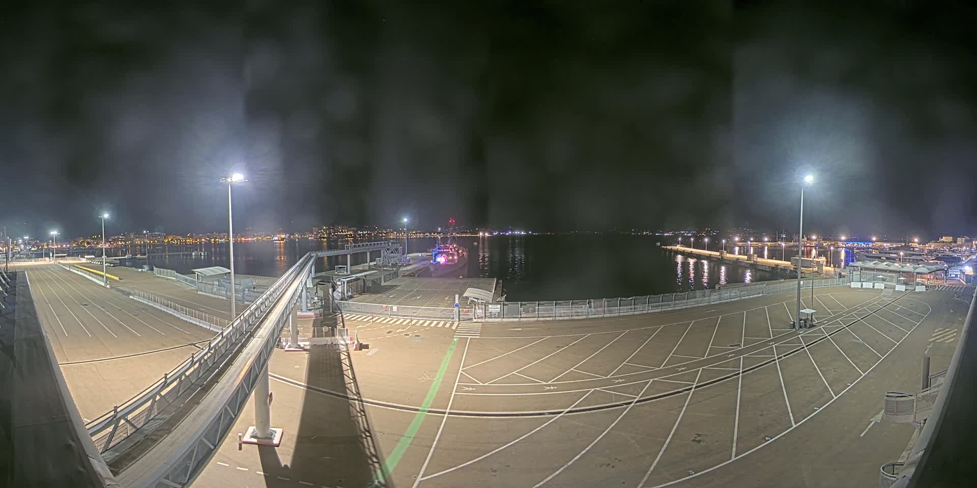 A nighttime view of an empty port area with a gangway leading to a docked vessel, under a hazy, dark sky.