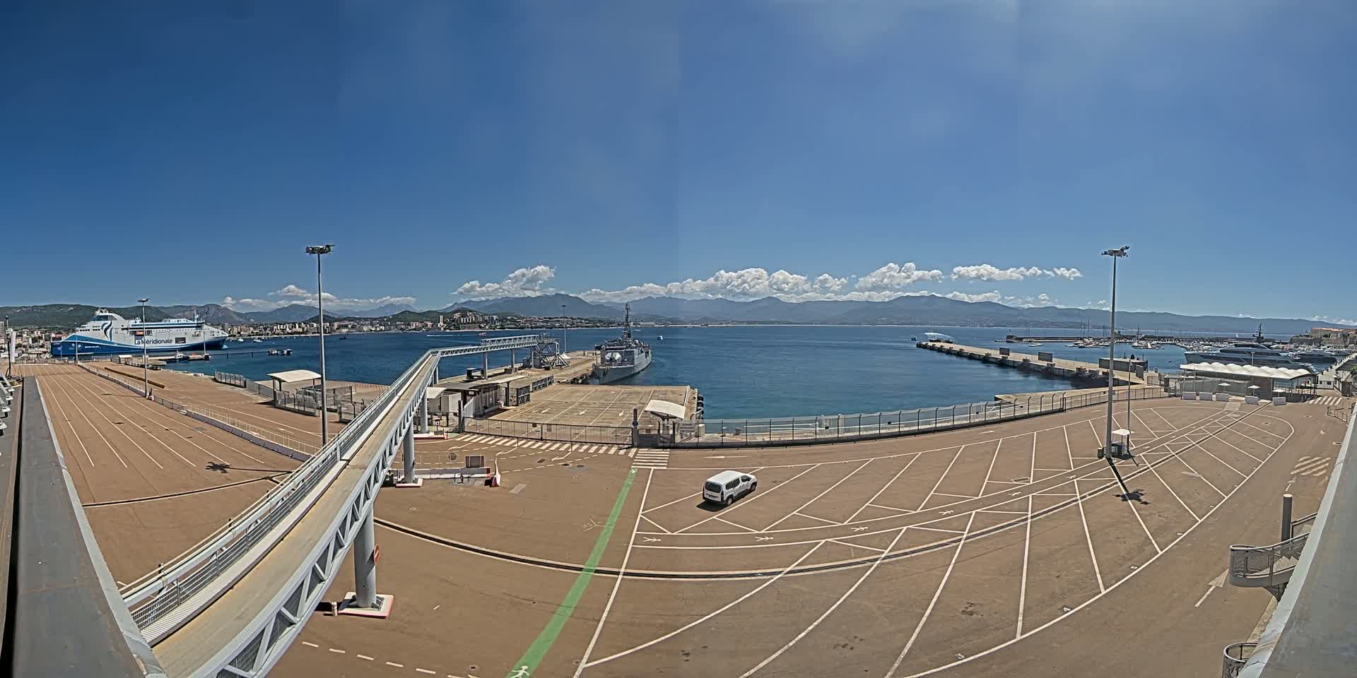 A ferry and a warship are docked at a port on a sunny day with partly cloudy skies and mountains visible in the distance.