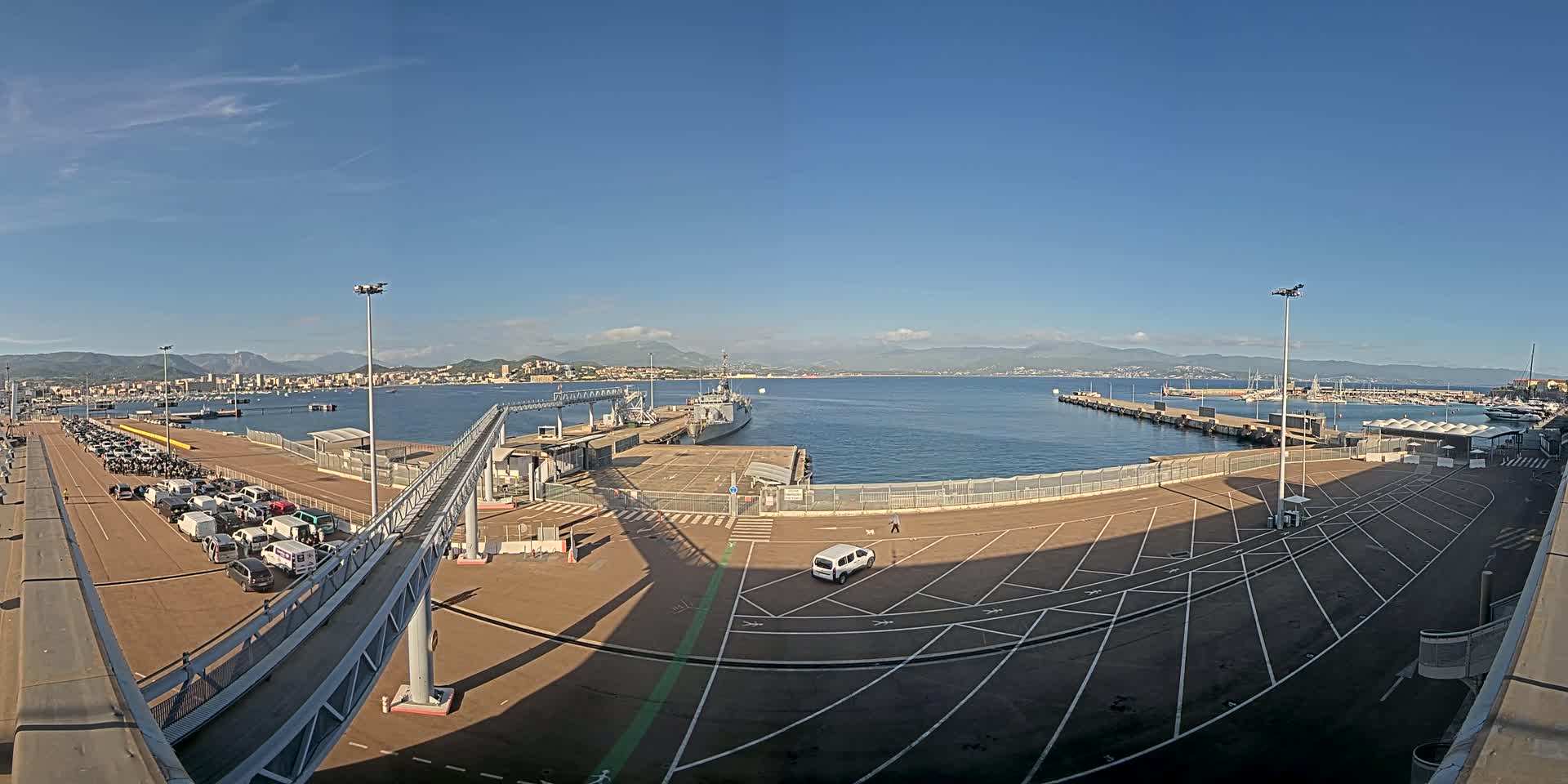 A ferry terminal with a large empty parking lot, a docked warship, and numerous vehicles parked near a gangway, all under a clear, sunny sky.