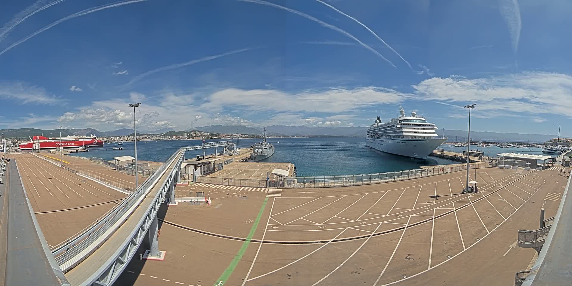 A cruise ship and a ferry are docked at a port on a sunny day with a partly cloudy sky and mountains in the background.