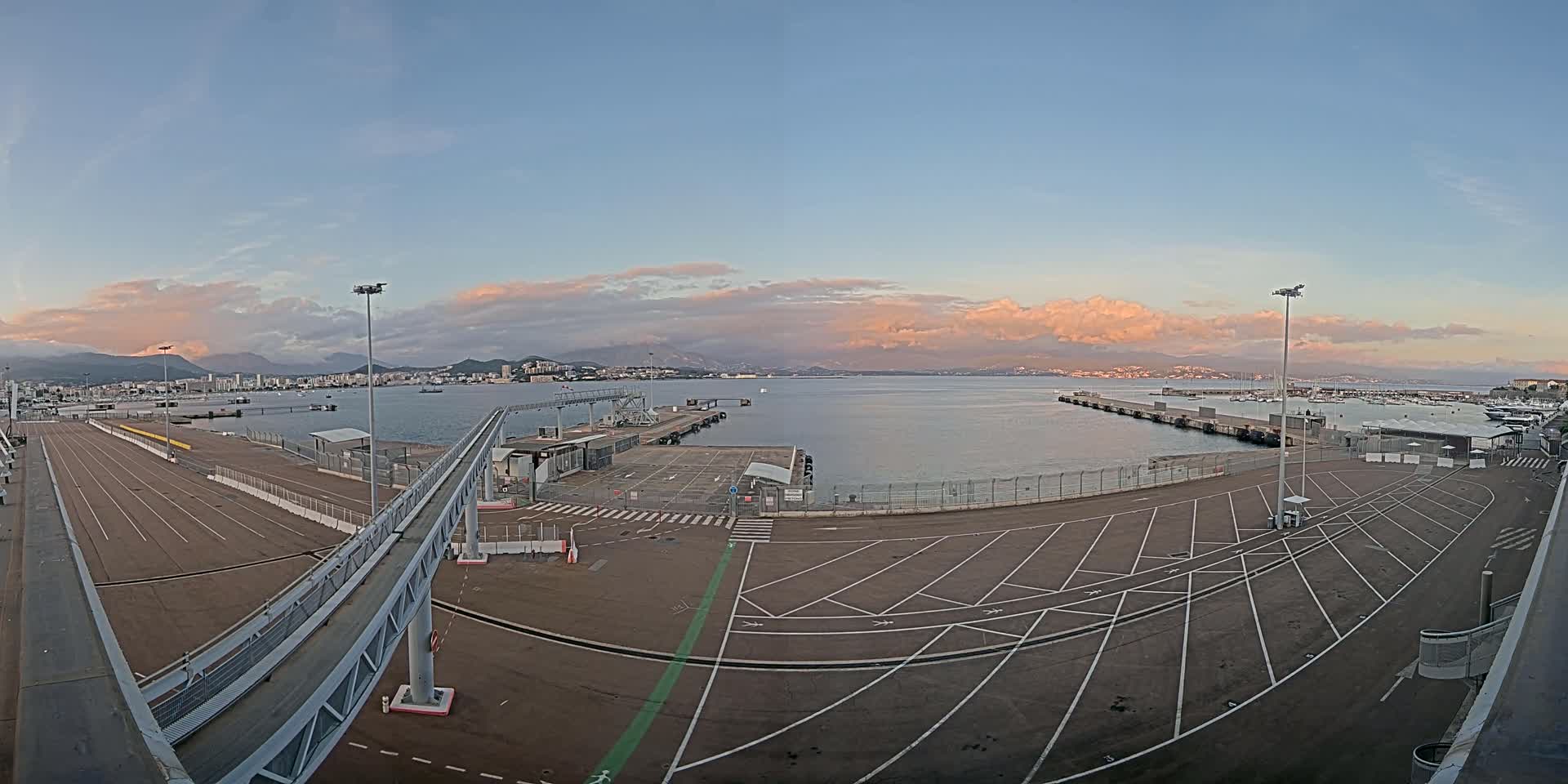 An empty port area with a long walkway leading to a dock, under a partly cloudy sunset sky with mountains in the distance.