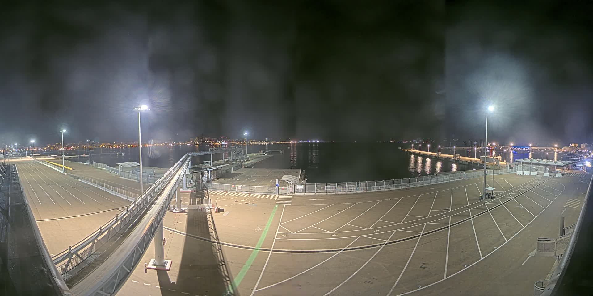 A nighttime view of a mostly empty port area with a gangway leading to a docked vessel, under a hazy, dark sky.