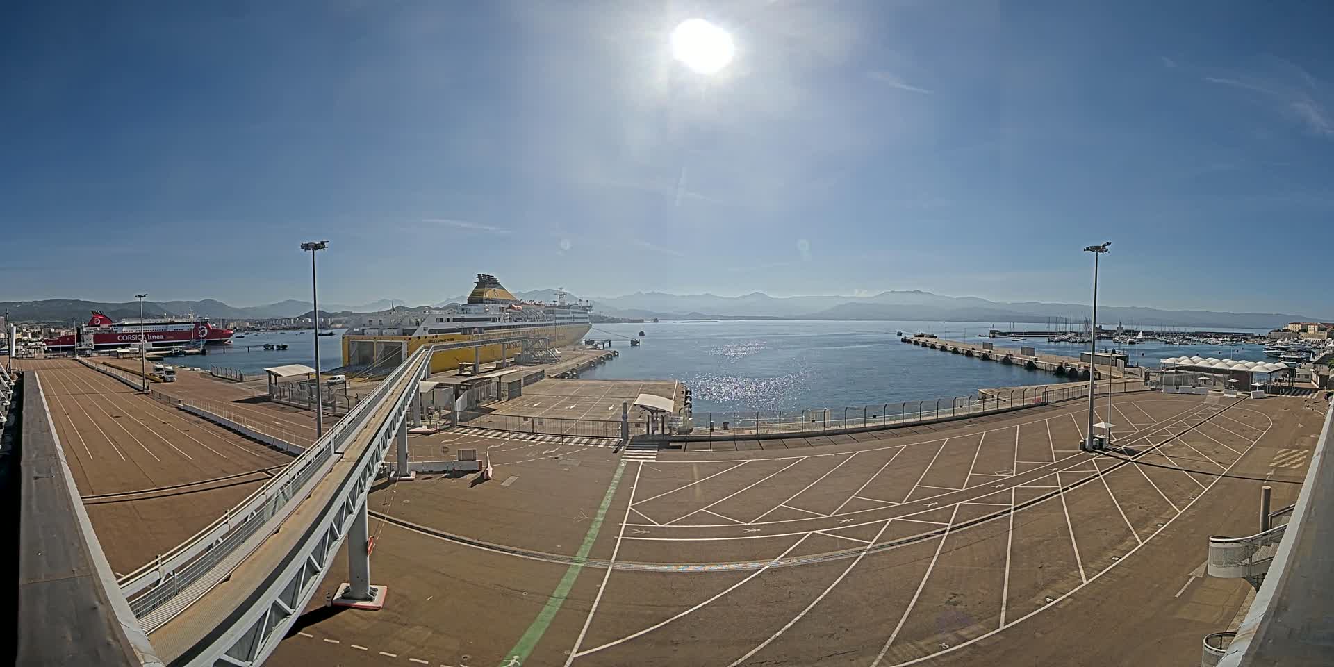 A large yellow ferry is docked at a port on a sunny day, with a smaller red ferry visible in the background and mountains in the distance.