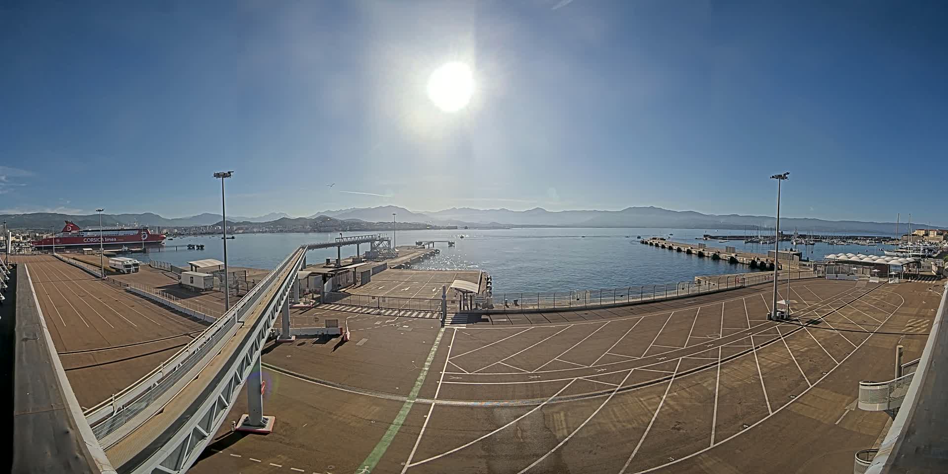 A sunny day at a harbor shows a large empty parking lot next to a dock with a passenger ferry and distant mountains visible across the calm water.