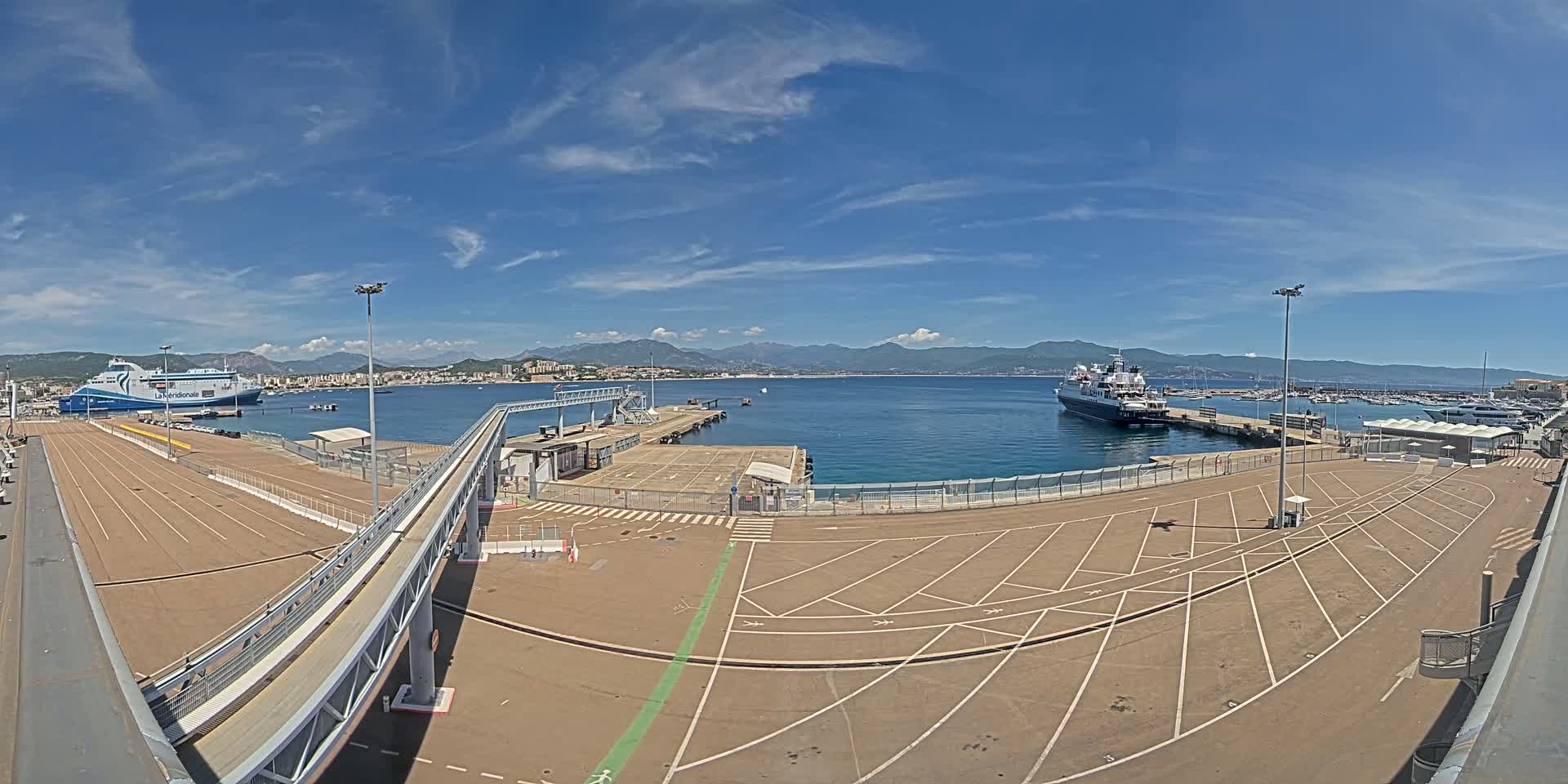 A ferry terminal on a sunny day with clear blue skies, featuring two large ships docked, one near the terminal and another further out in the harbor, with a large empty parking lot in the foreground and a pedestrian walkway leading to the docked ferry.