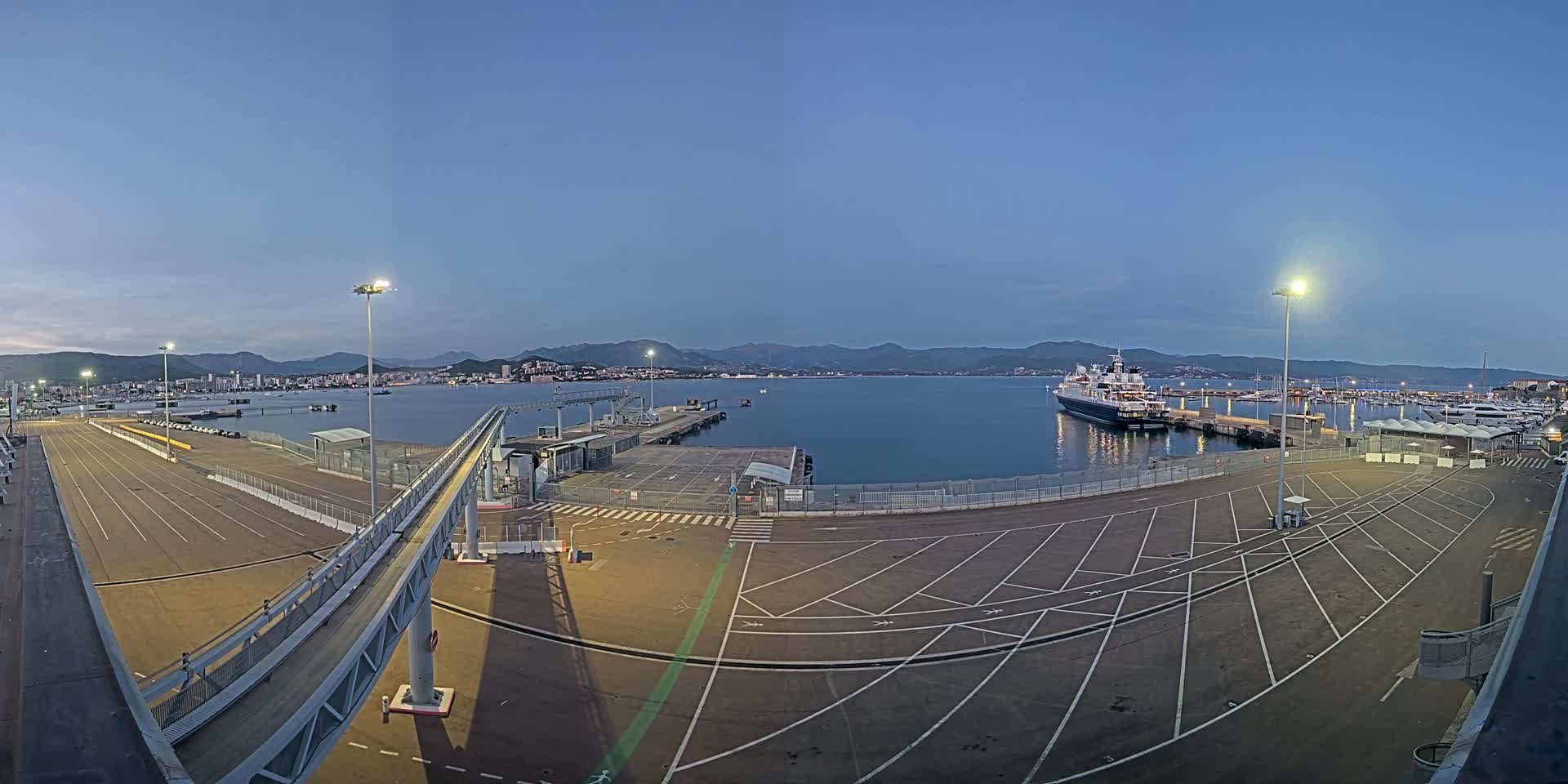 An empty port at dusk, with a large cruise ship and numerous smaller boats moored in the calm water, mountains visible in the distance under a clear sky.