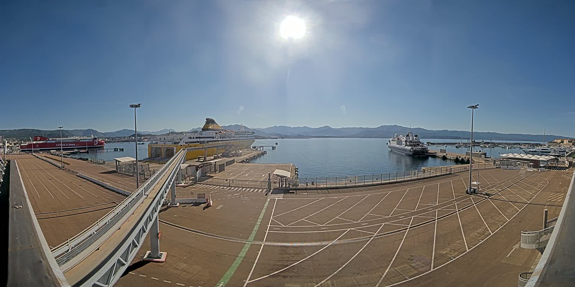 A sunny day at a port shows two large ferries docked, along with other smaller boats, and a large empty parking lot.
