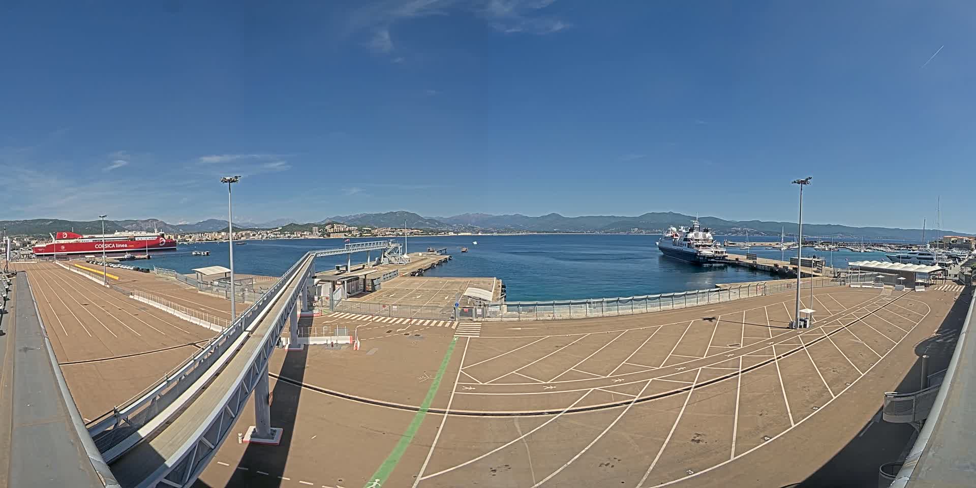 A ferry terminal with a large empty parking lot, a passenger gangway, and two ships in a harbor on a sunny day with clear blue skies and distant mountains.