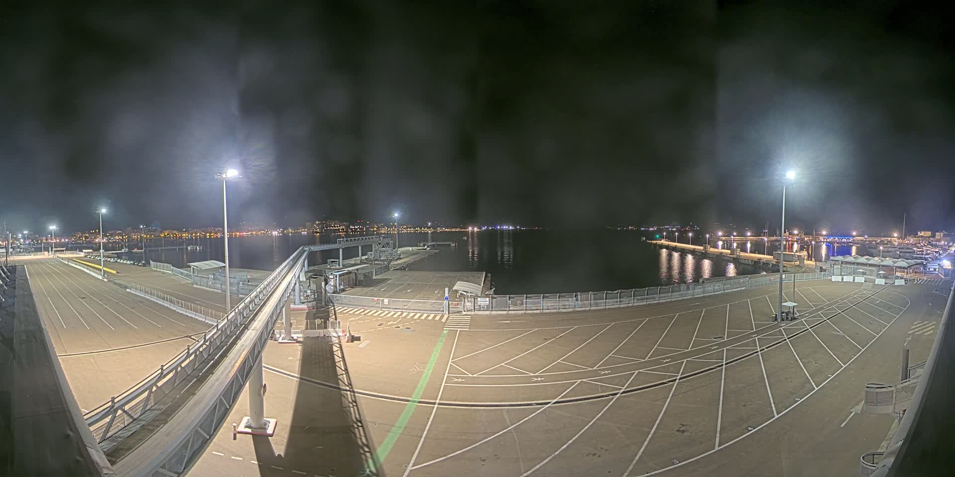 A nighttime view of a mostly empty port area with a long, elevated walkway, under a hazy sky.