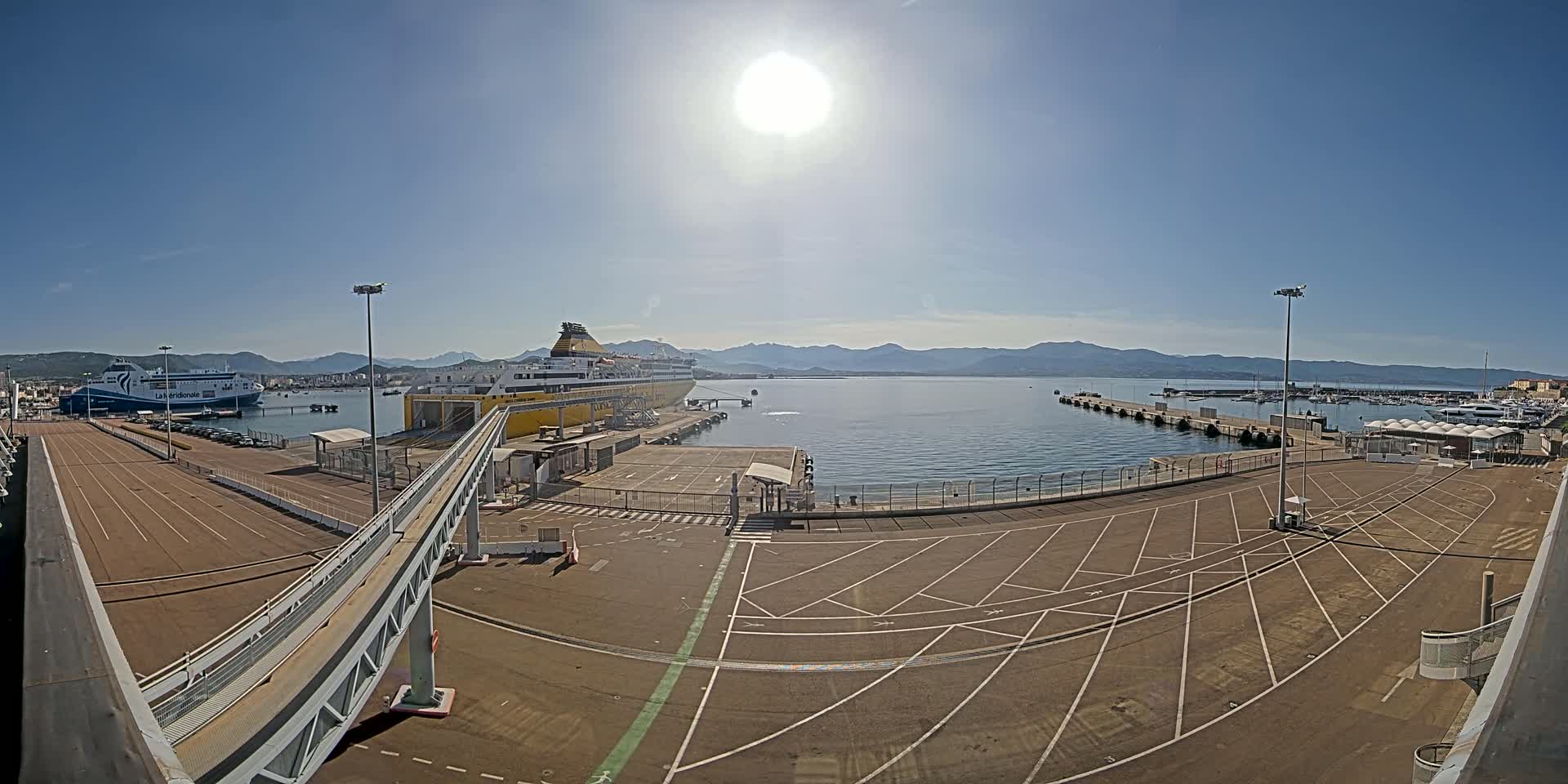 A large yellow ferry is docked at a port on a sunny day, with a smaller ferry and many smaller boats visible in the harbor, and mountains visible in the distance.
