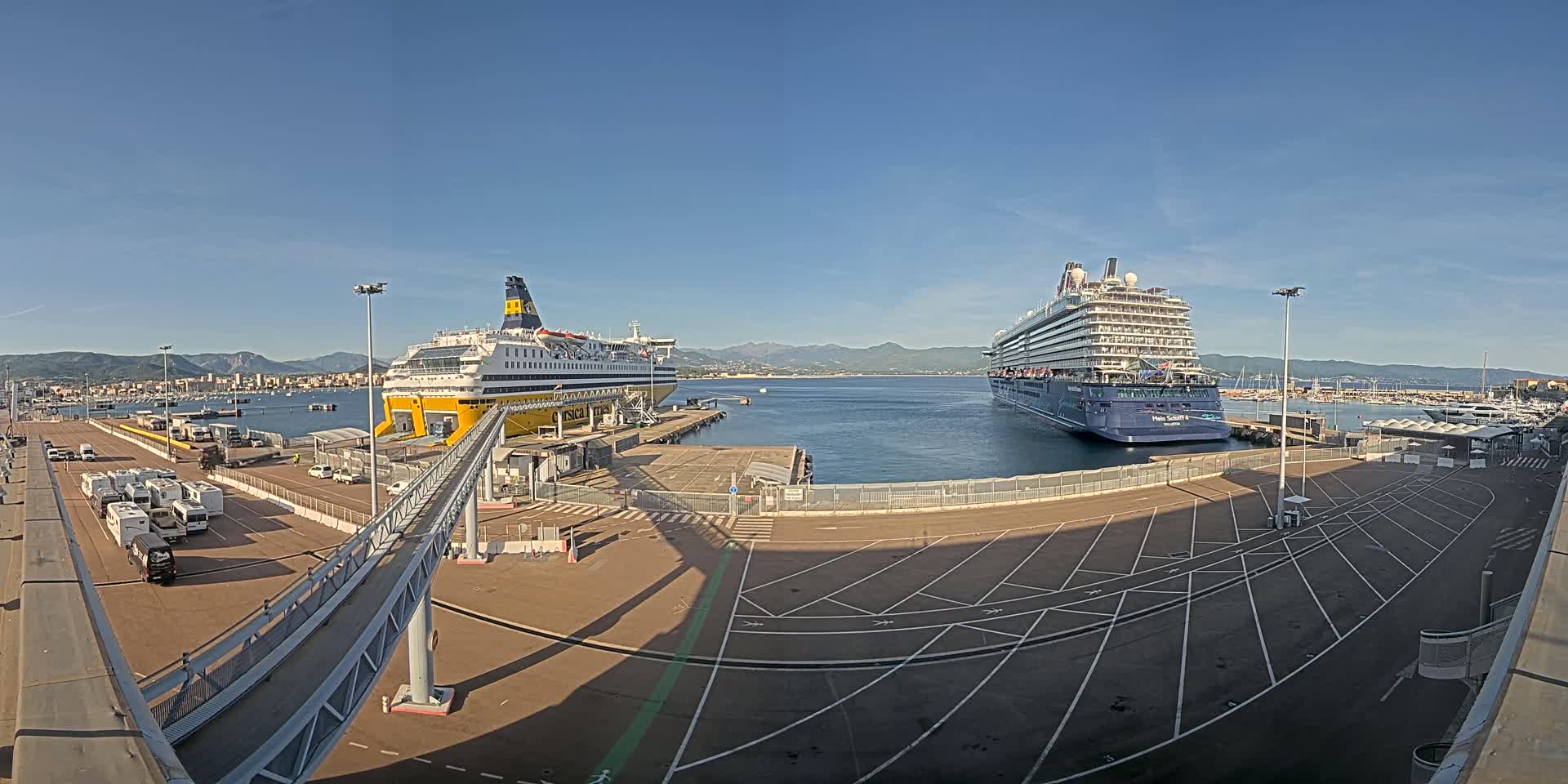 A large ferry and a cruise ship are docked at a port on a sunny day, with mountains visible in the distance.
