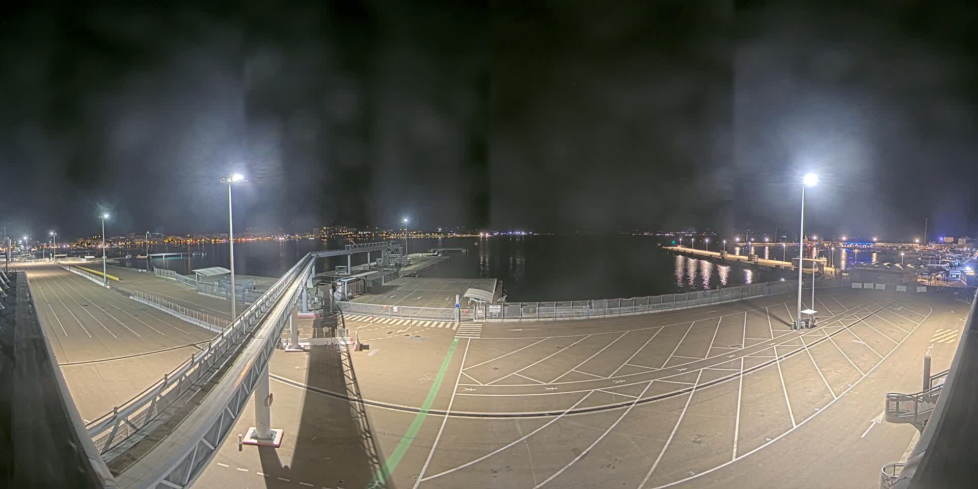 A nighttime view of an empty ferry terminal and parking lot overlooking a calm body of water with a city skyline in the distance.