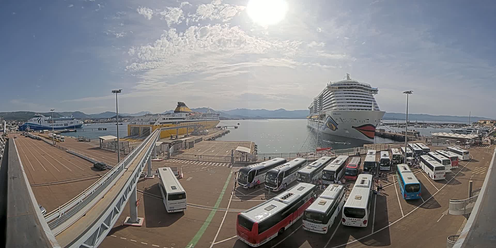A sunny day at a port shows two large ships docked, one ferry and one cruise ship, with numerous buses parked nearby.