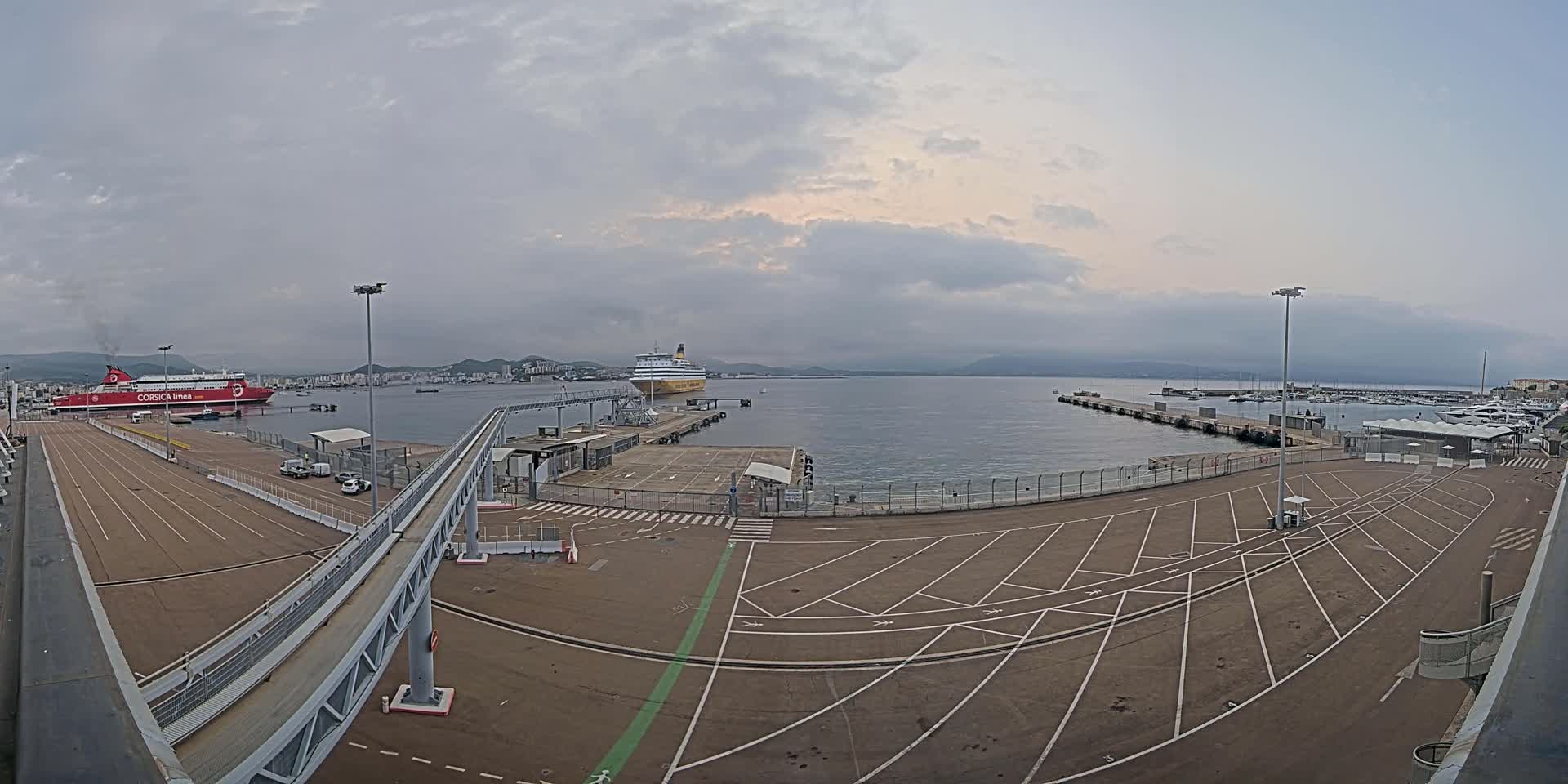 A ferry terminal with two ferries docked, one red and one yellow, under a mostly cloudy sky.