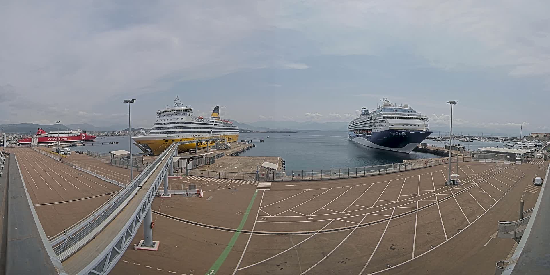 A large cruise ship and a ferry are docked at a port under a mostly cloudy sky.