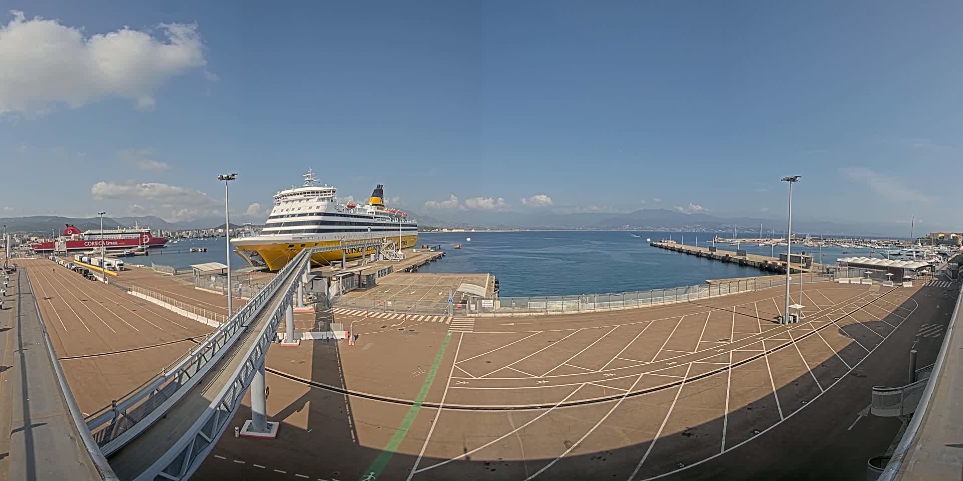 A large ferry is docked at a port on a sunny day with calm water and mountains visible in the distance.