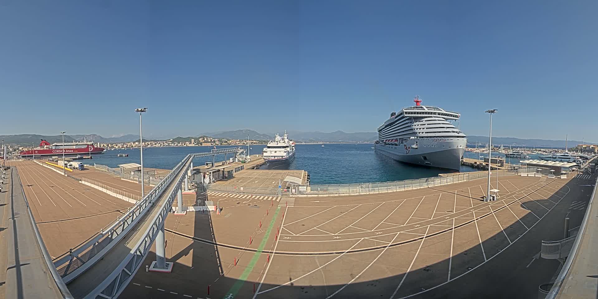 A cruise ship and a ferry are docked at a port on a sunny day, with mountains visible in the distance.