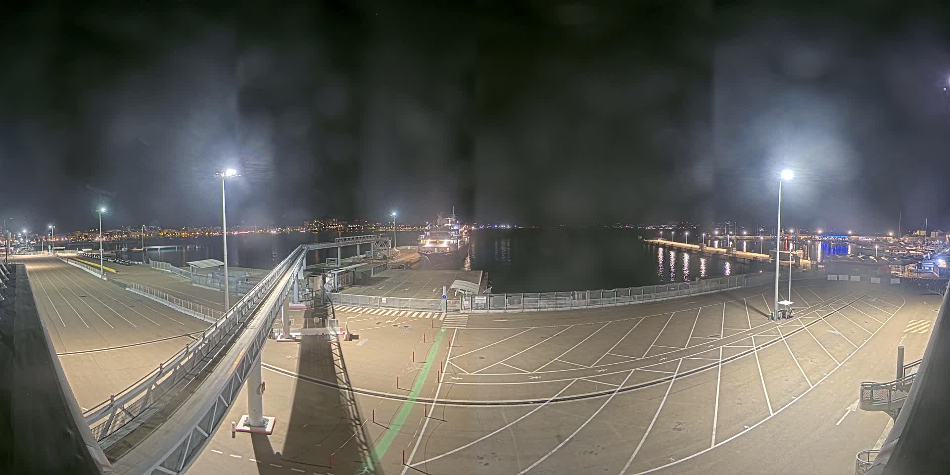A nighttime view of a mostly empty port with a ship docked, illuminated by several tall lights under a hazy, dark sky.