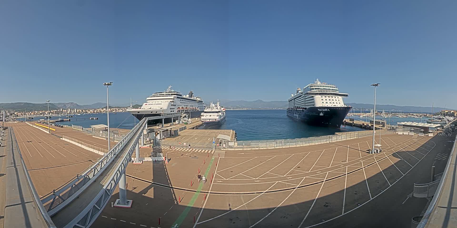 Three cruise ships are docked at a port on a sunny day, with mountains visible in the distance.