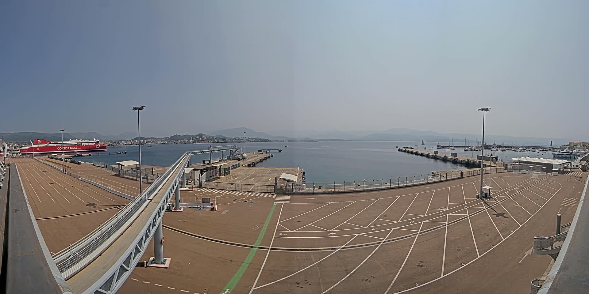 A ferry is docked at a port on a clear, sunny day, with a large empty parking lot and a pedestrian walkway leading to the ship.