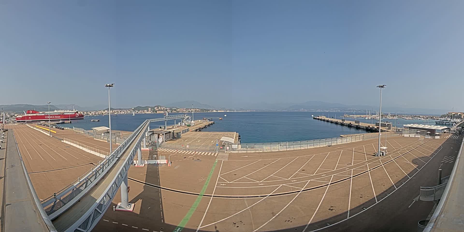 A large, empty parking lot at a port, a red ferry docked nearby, and a clear, sunny day with calm water and distant mountains visible across the bay.
