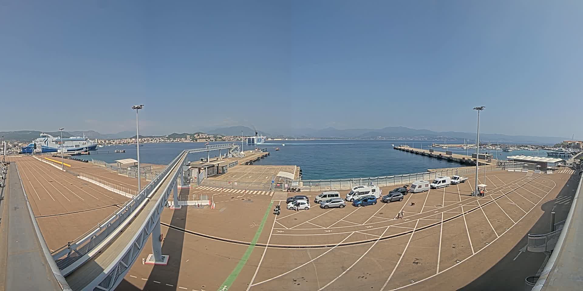 A ferry terminal on a sunny day with several vehicles parked in a designated area, a docked ferry visible in the background, and a walkway leading to the pier.
