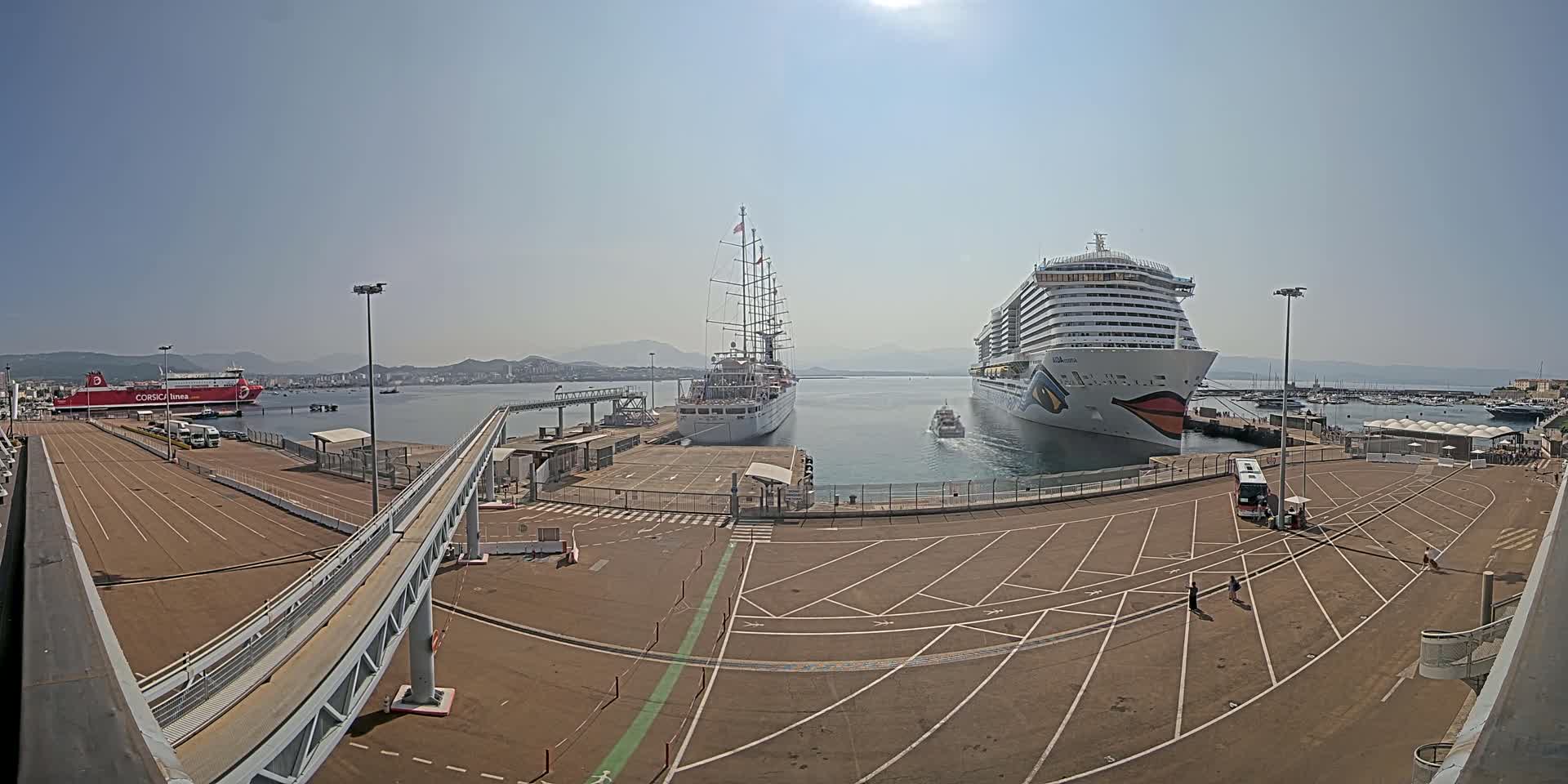 A cruise ship and a sailing ship are docked at a port on a sunny day, with a ferry and other smaller boats visible in the background.