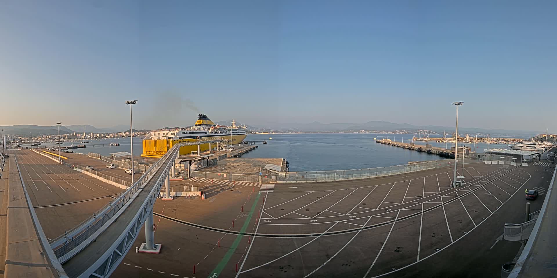 A large yellow ferry is docked at a port on a calm, sunny day, with a wide empty parking lot and a pedestrian walkway leading to the ship.
