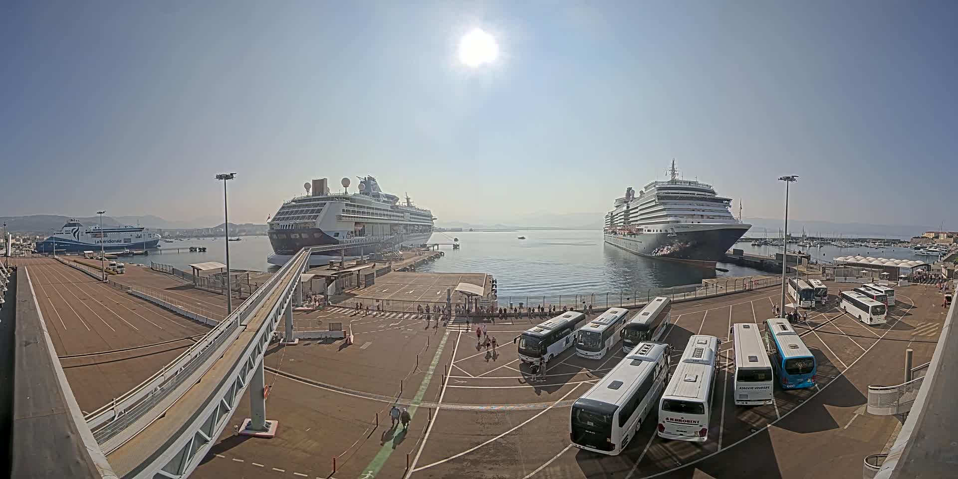 Two large cruise ships and a ferry are docked at a port on a sunny day, with numerous buses parked nearby.