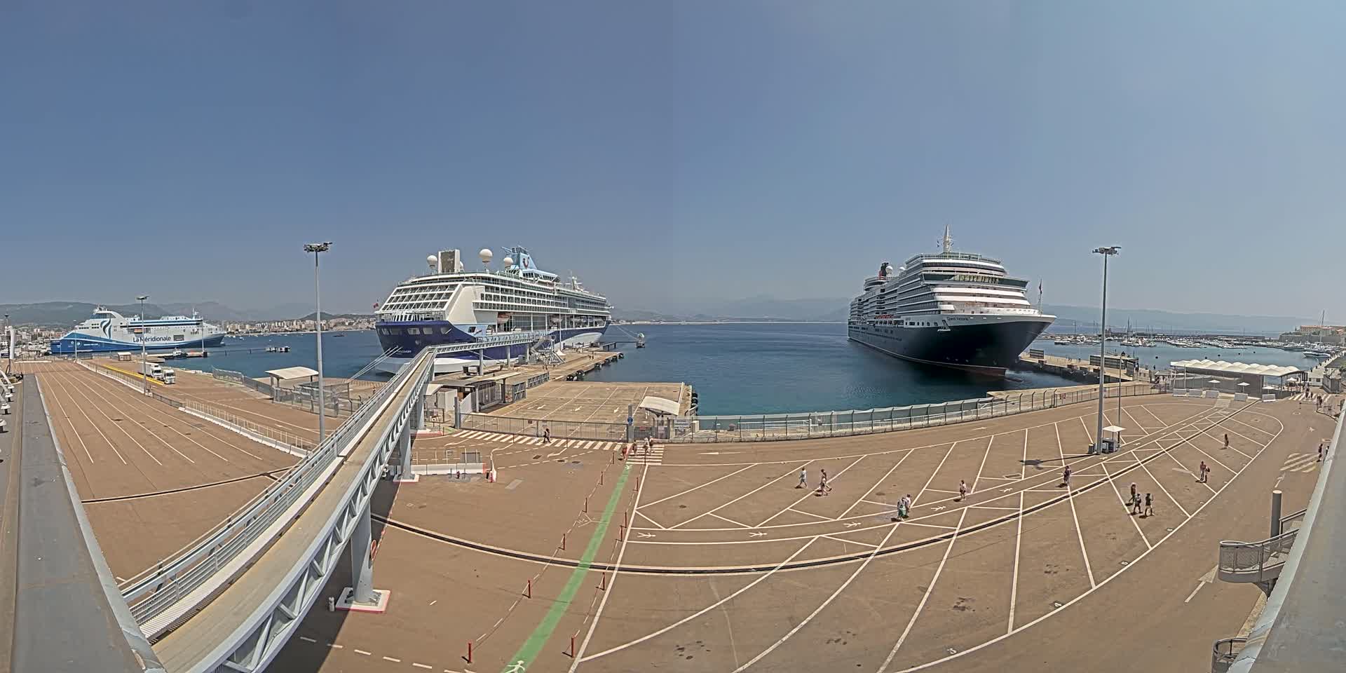 Two large cruise ships are docked at a port on a sunny day, with a smaller ferry visible in the background.