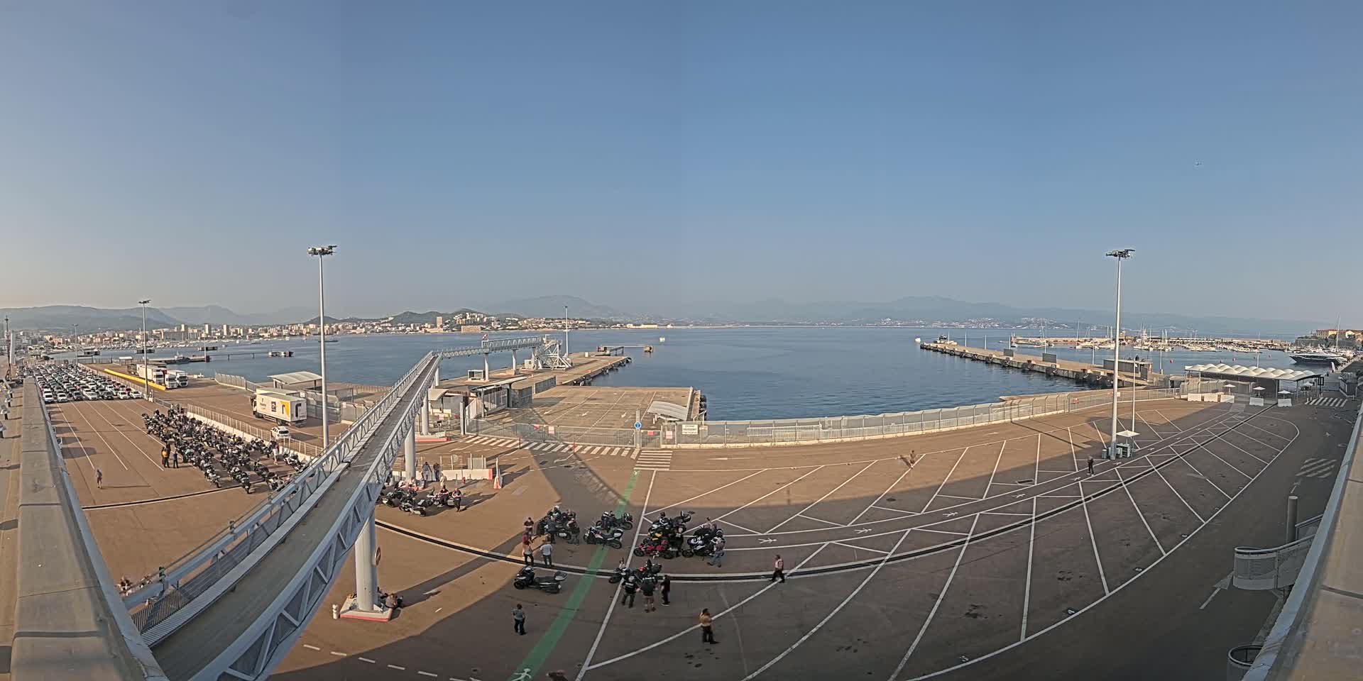 A large empty port area with parked cars and motorcycles, a walkway leading to a dock, and a calm sea with a distant city and mountains under a clear blue sky.