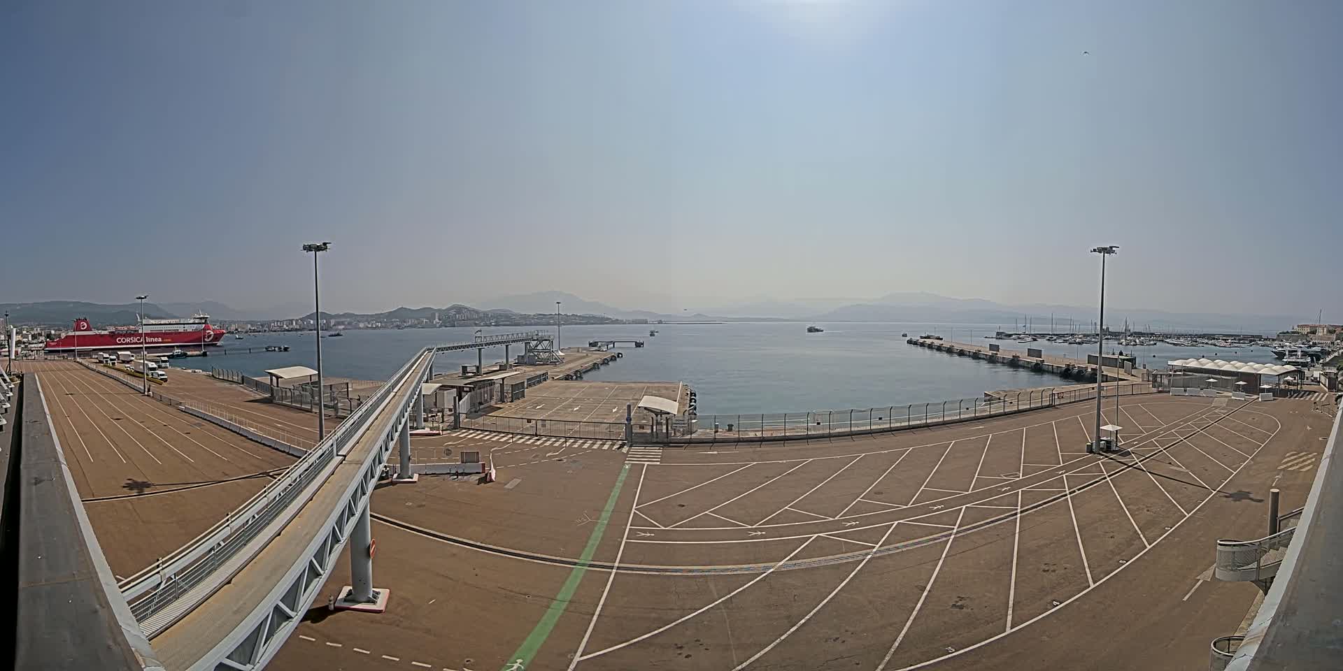 A large empty parking lot at a ferry terminal overlooks a calm body of water with a docked ferry and numerous boats under a clear, sunny sky.