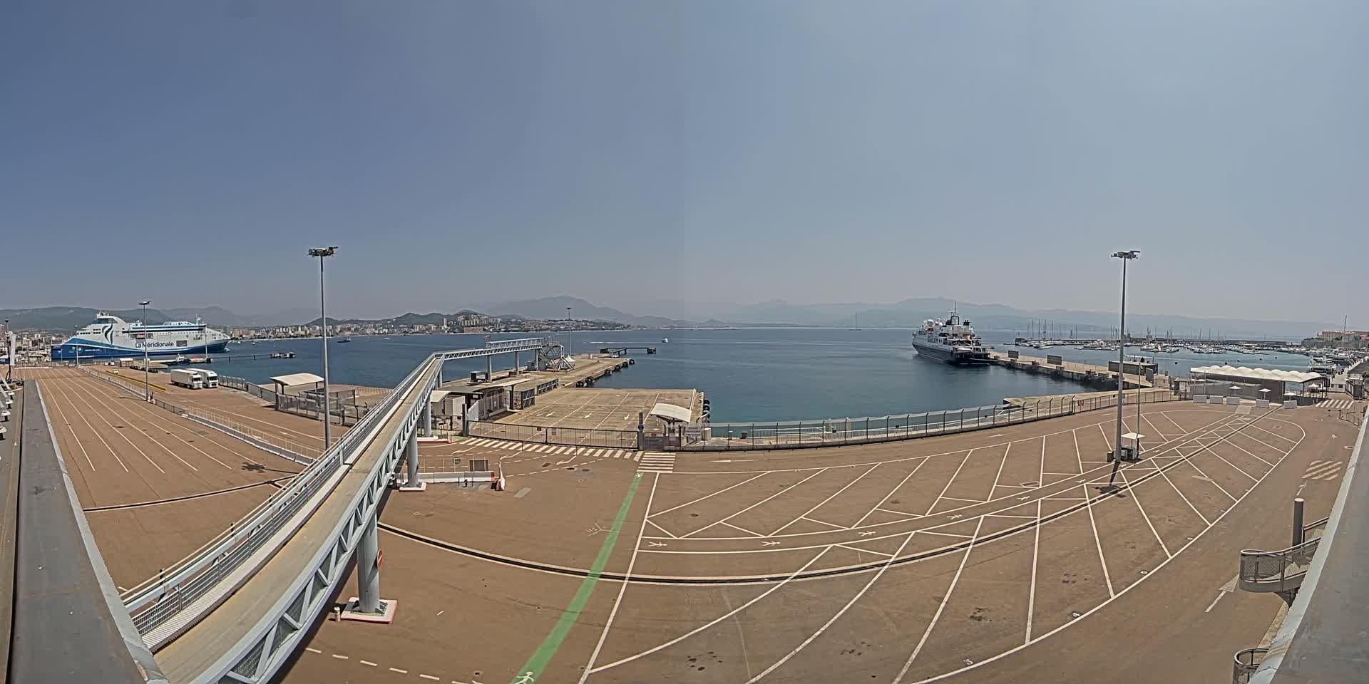 A ferry terminal with empty parking lots and two ships in the harbor on a clear, sunny day.