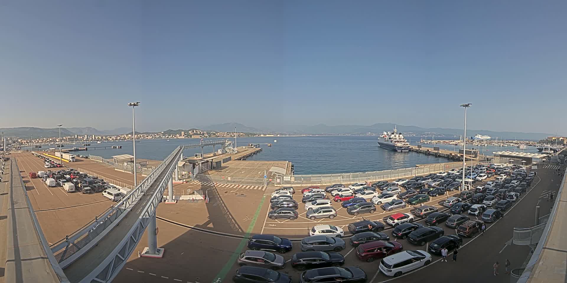 A ferry terminal with numerous cars parked in lots, a gangway, and a large body of water with a ferry and distant coastline under sunny skies.